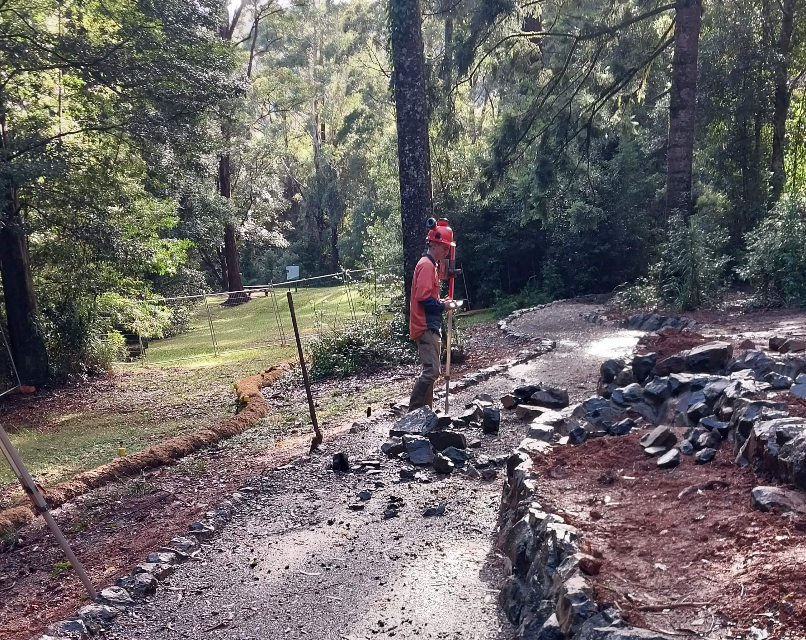 A man in high-vis clothing and a safety helmet works on a rock-lined path against a rainforest backdrop