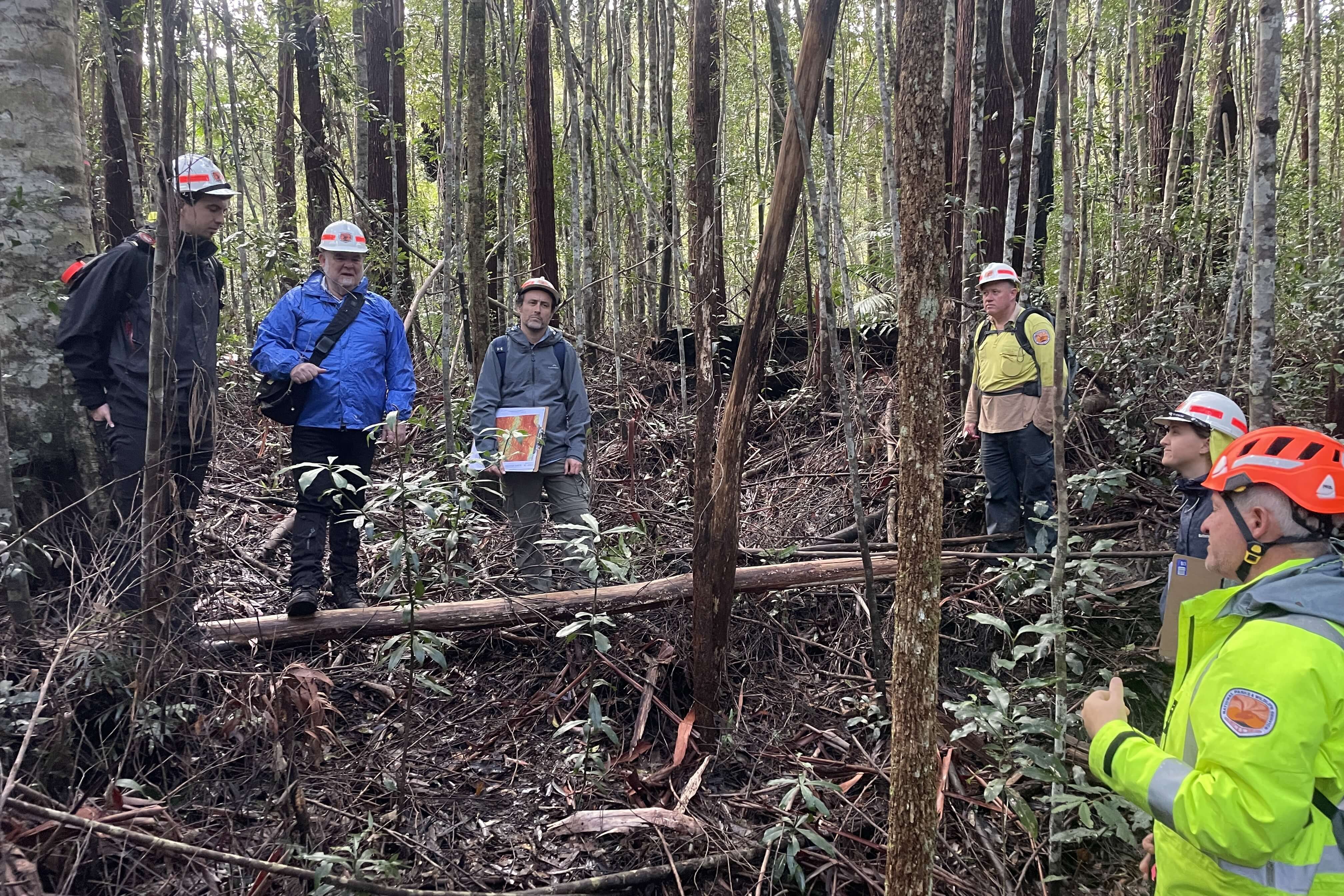 Six men in safety helmets and warm clothing stand in a circle in dense forest