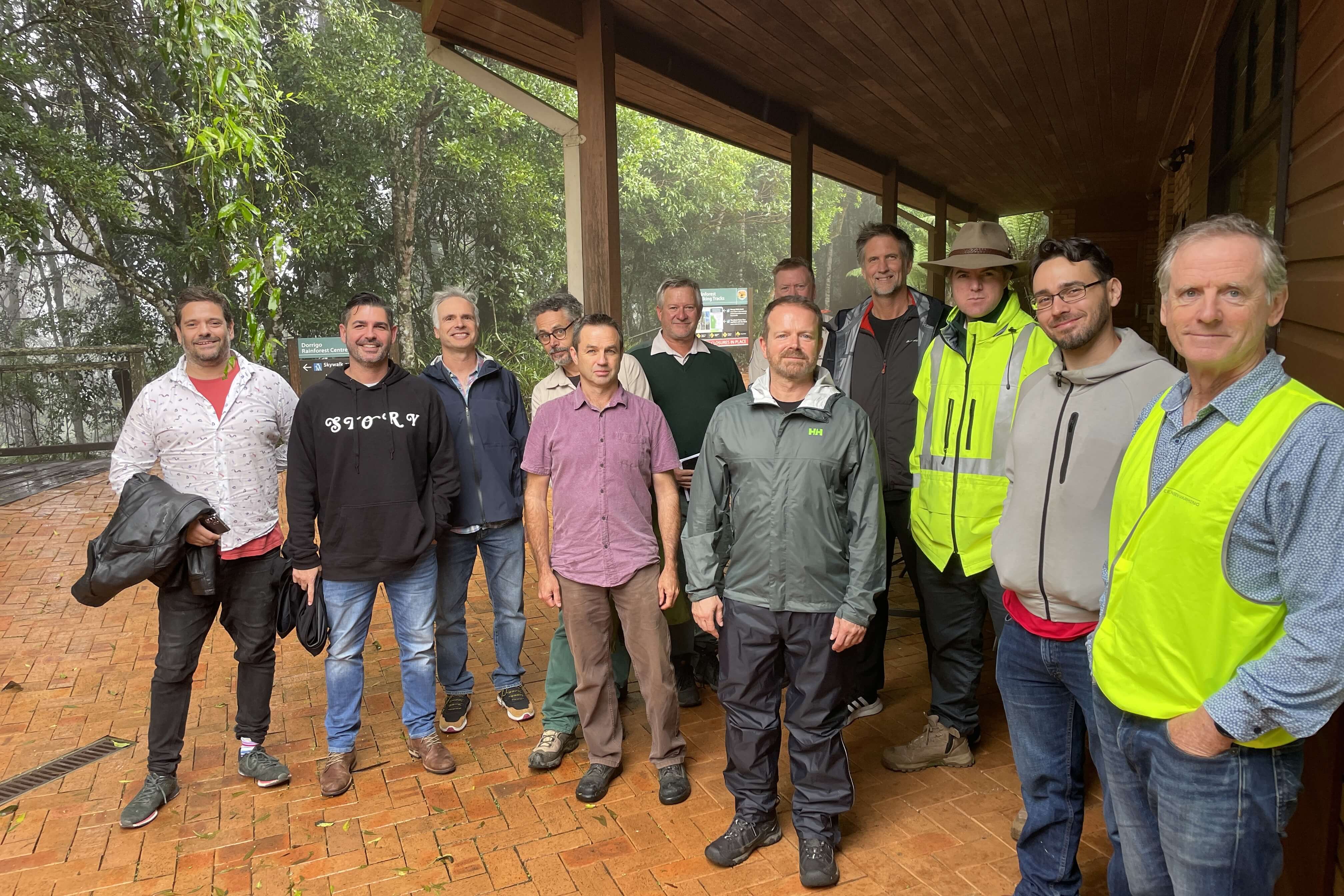 Twelve men smile and pose for the camera against a backdrop of rainforest and a low-set building.