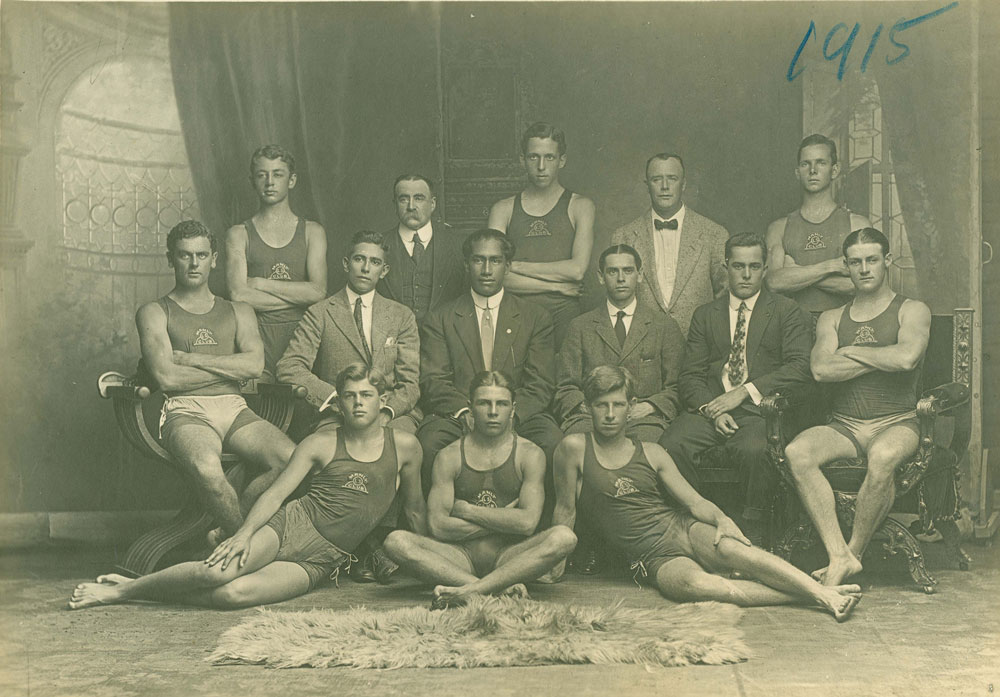 Formally posed sepia portrait of Duke Kahanamoku, seated centre wearing a suit, surrounded by Manly Surf Life Saving Club Members taken in 1915 