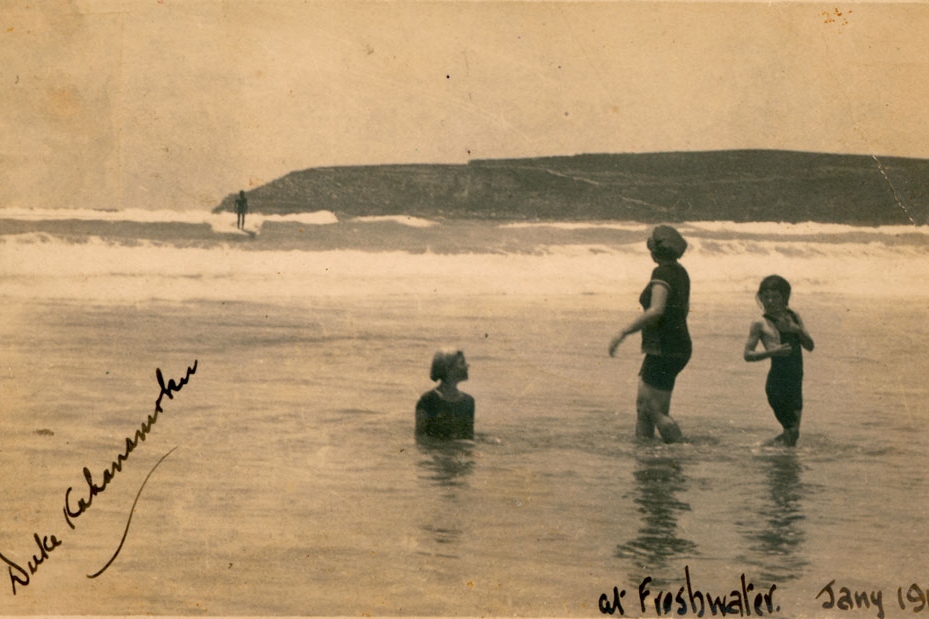 A sepia photo showing a man surfing. Three swimmers in the foreground look out to the man on the waves. The photographer has written on the front of the photo 'Duke Kahanamoku, 1915'
