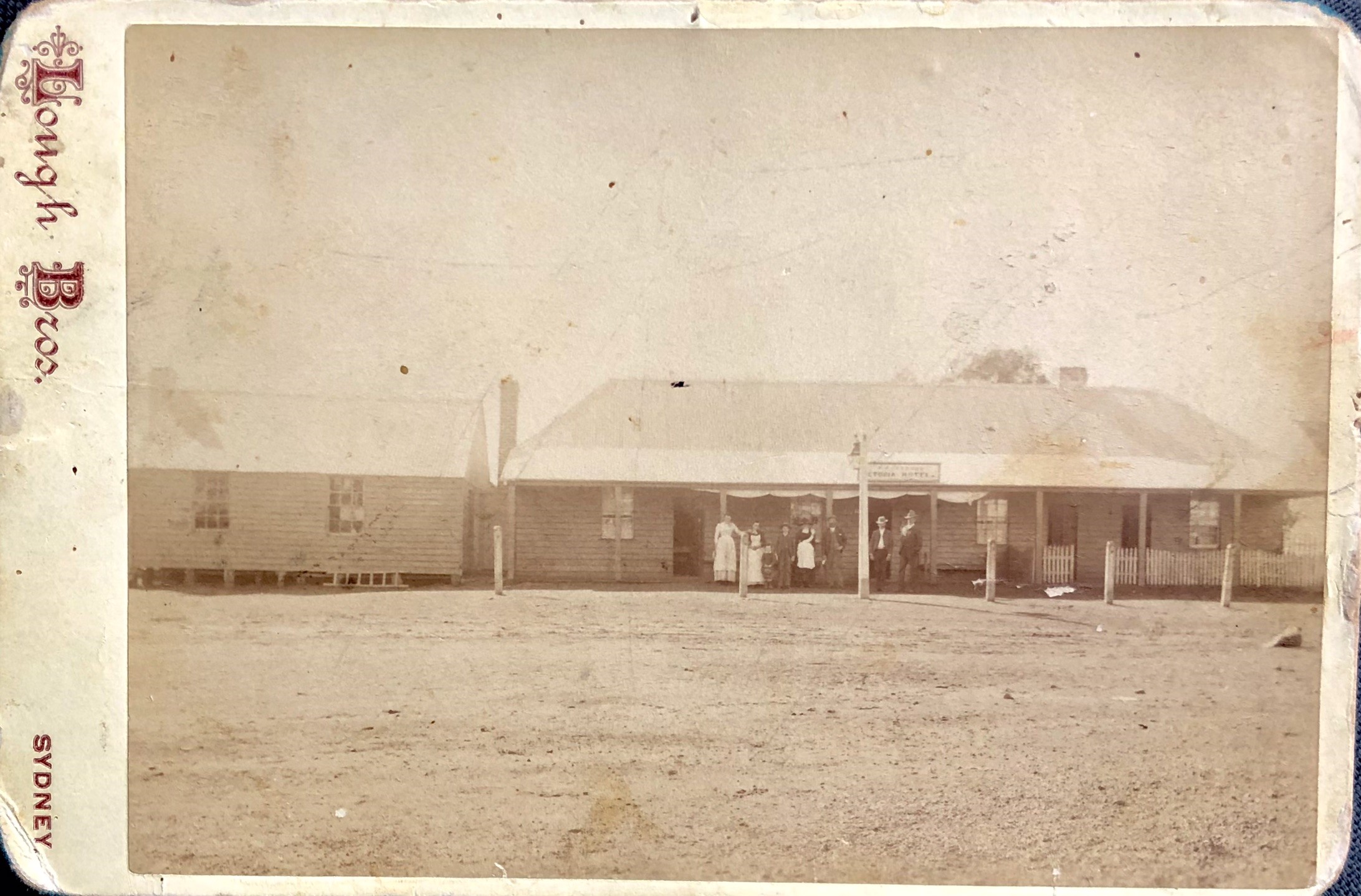 A sepia photograph of the Victoria Hotel, now known as the Old Vic. A group of seven people are standing under the front verandah looking to the camera.