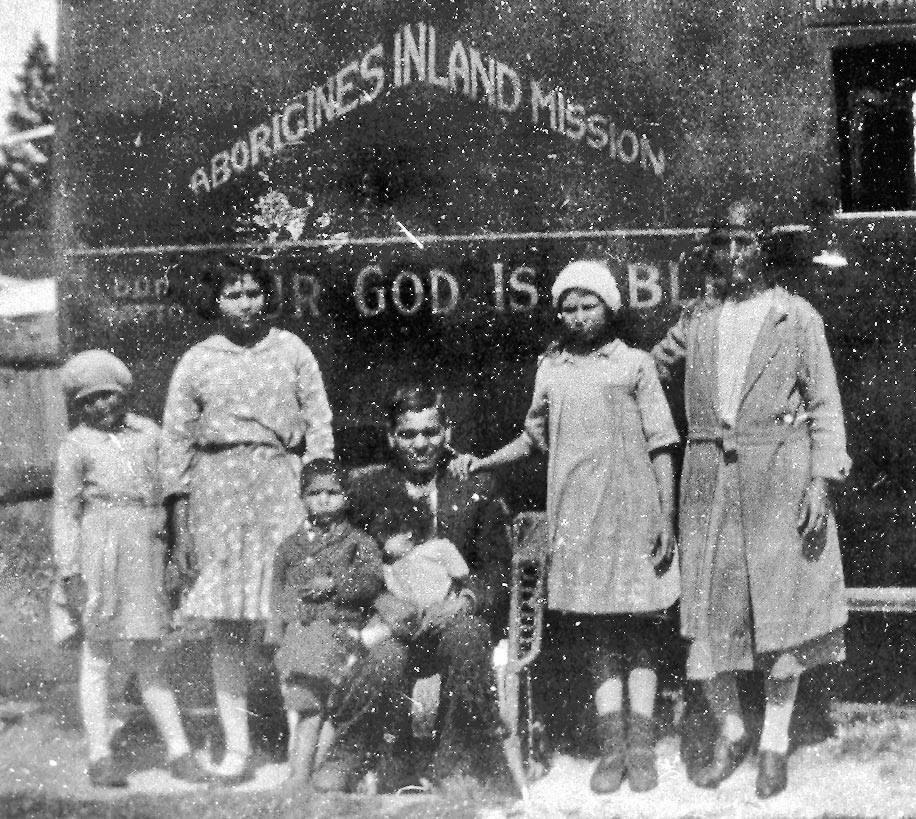 Black and white photograph of Emma Jane Callaghan with her family in Armidale. Seven people are pictured: two girls standing on the left and two girls on the right. In the center, a man is seated on a chair holding a toddler. To his right (photo’s left), a young boy stands beside him. In the background, a sign reads 'Aboriginal Inland Mission,' indicating the setting’s historical and cultural context.