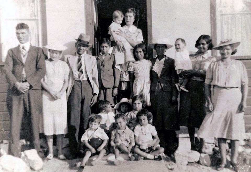 Black and white photograph of Emma Jane Callaghan with her extended family in front of her home on Tasman Street, La Perouse. A total of 17 people are pictured. Six young children are seated on the doorstep, while teenagers and adults stand behind them. Several adults are holding babies, capturing a multigenerational family moment outside a modest home.