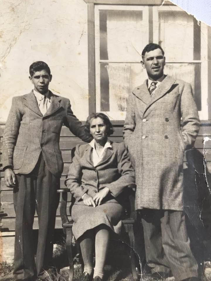 Black and white photo of a woman sitting down and 2 men standing on either side of her, with a building behind them