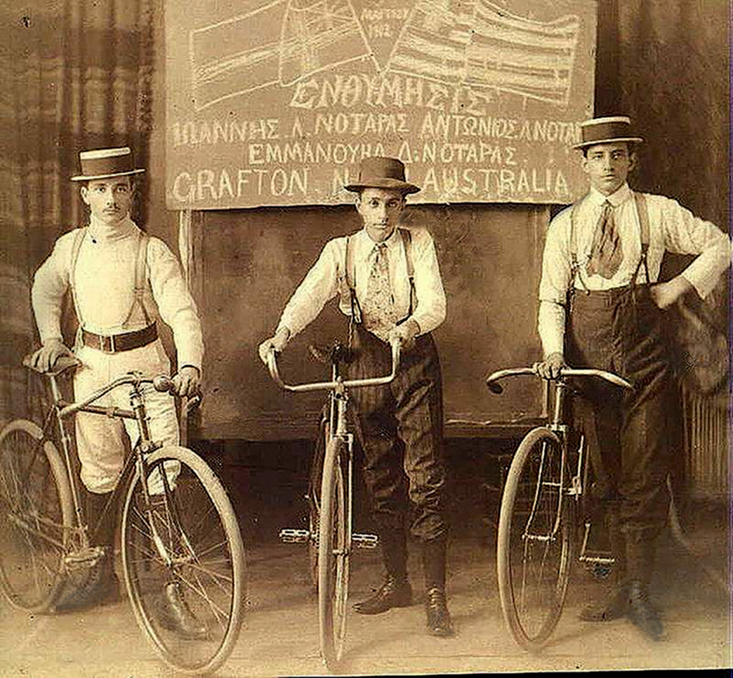 epia-toned photograph taken in Grafton, 1912, featuring Emmanuel Dimitrios Notaras and his first cousins Anthony and John Lambrinos Notaras. The three young men are standing side by side, each holding a bicycle. They are dressed in early 20th-century attire: wide-brimmed hats, white shirts or tops, trousers held up by suspenders, and black boots. Behind them, a sign reads their names along with “Grafton, Australia".