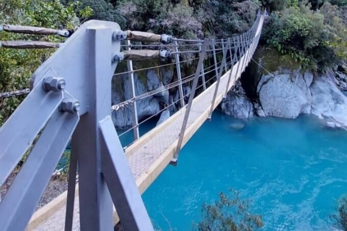 A narrow suspension bridge over a creek with clear blue water, in a forest