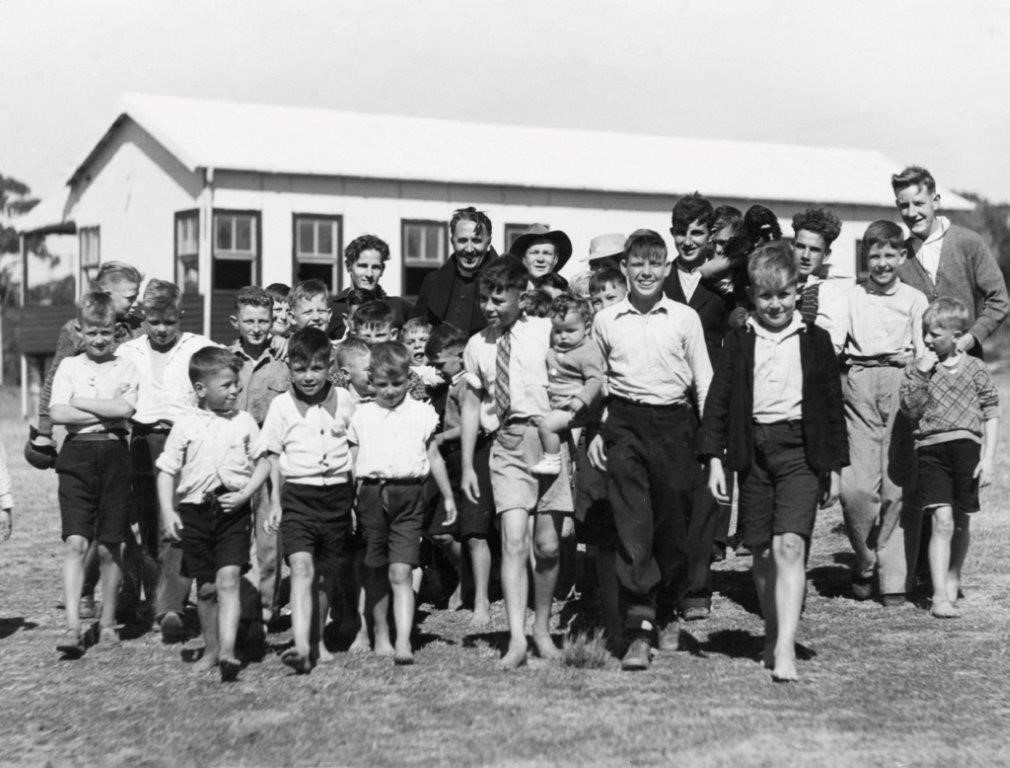A young Father Dunlea with a large group of boys of all ages standing in front of a building at Boys Town in Engadine, now known as the Dunlea Centre.