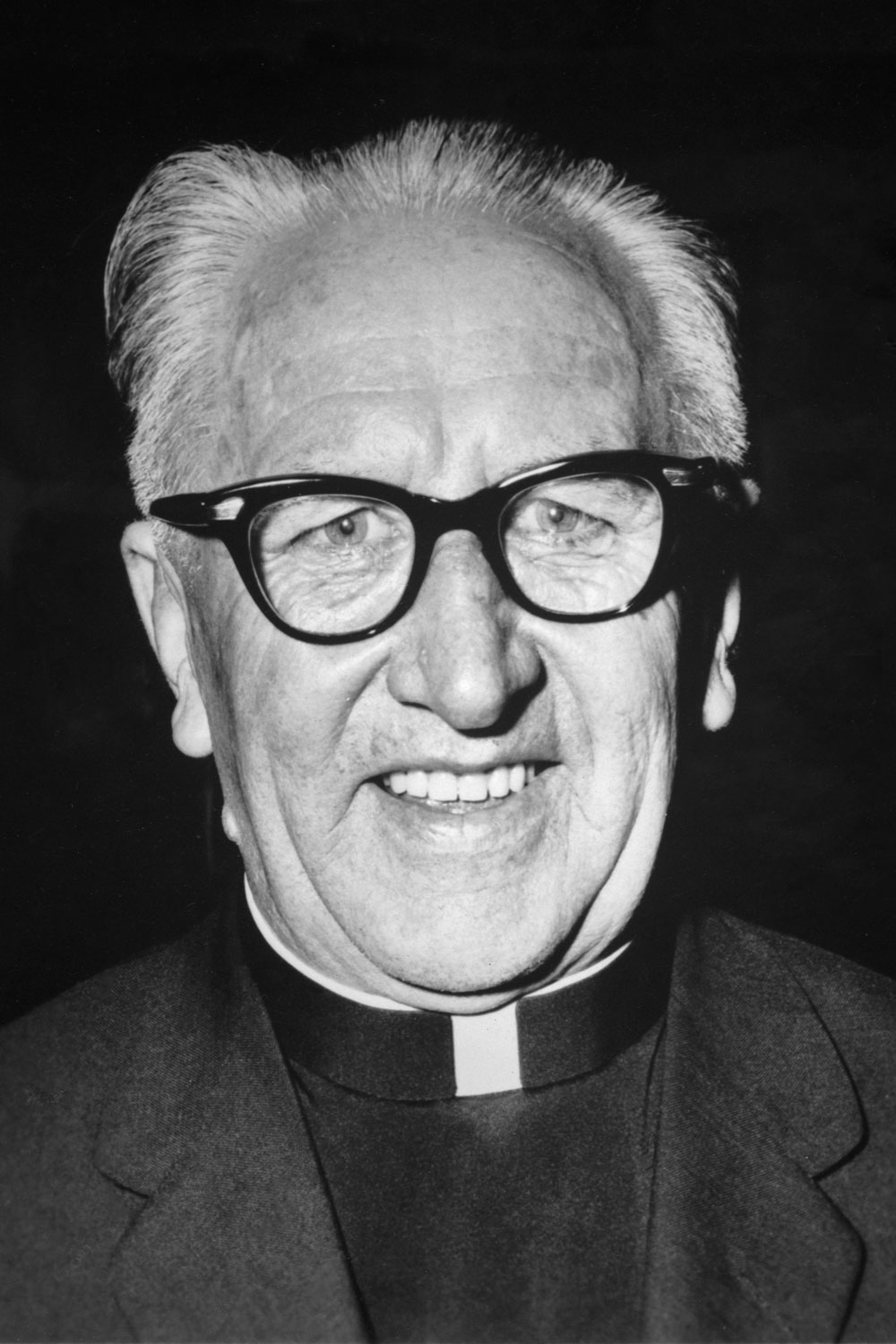A closeup black and white photograph of a smiling Father Thomas Dunlea, he wears glasses and priest’s vestments.
