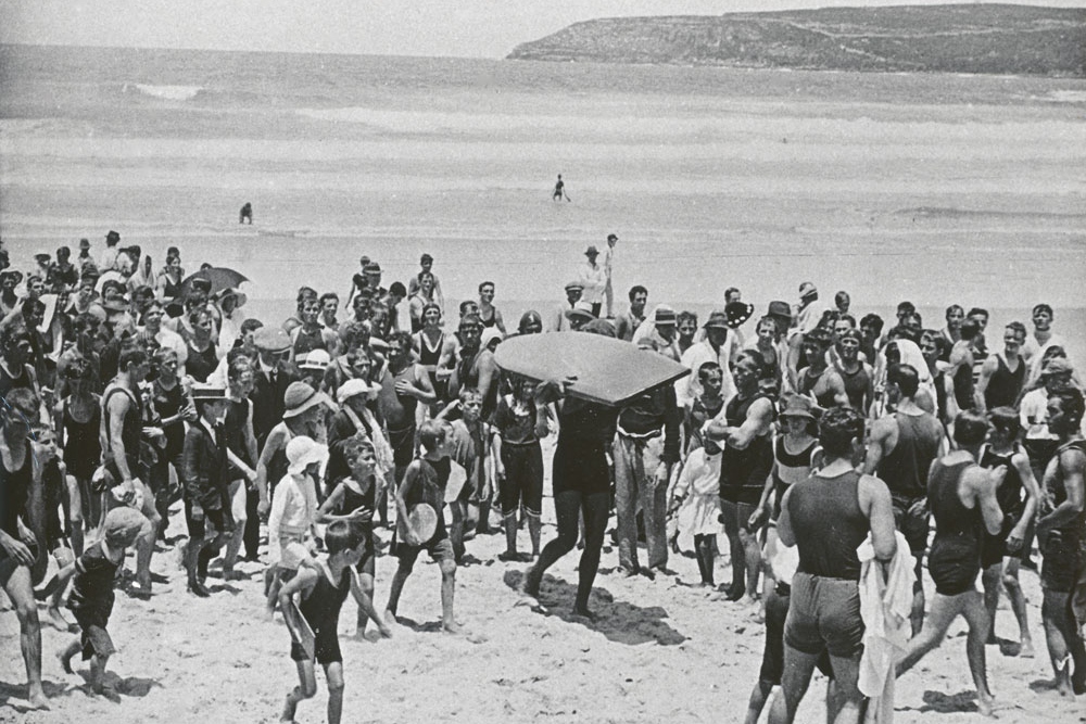 A black and white photo showing a crowd surrounding a man carrying a large surfboard on a beach