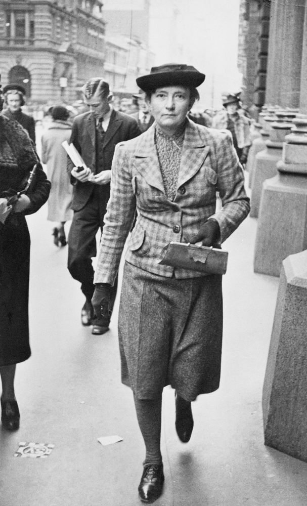Black and white image of Grace Cossington Smith walking down the street with other people walking in the background. She is holding a small purse while wearing shoes, stockings, a skirt, a plaid blazer, gloves and a hat.