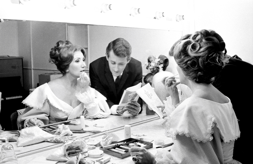 Black and white image of June Bronhill applying her make-up in front of the dressing room mirror with her manager. June is wearing a dress and the man is wearing a suit.