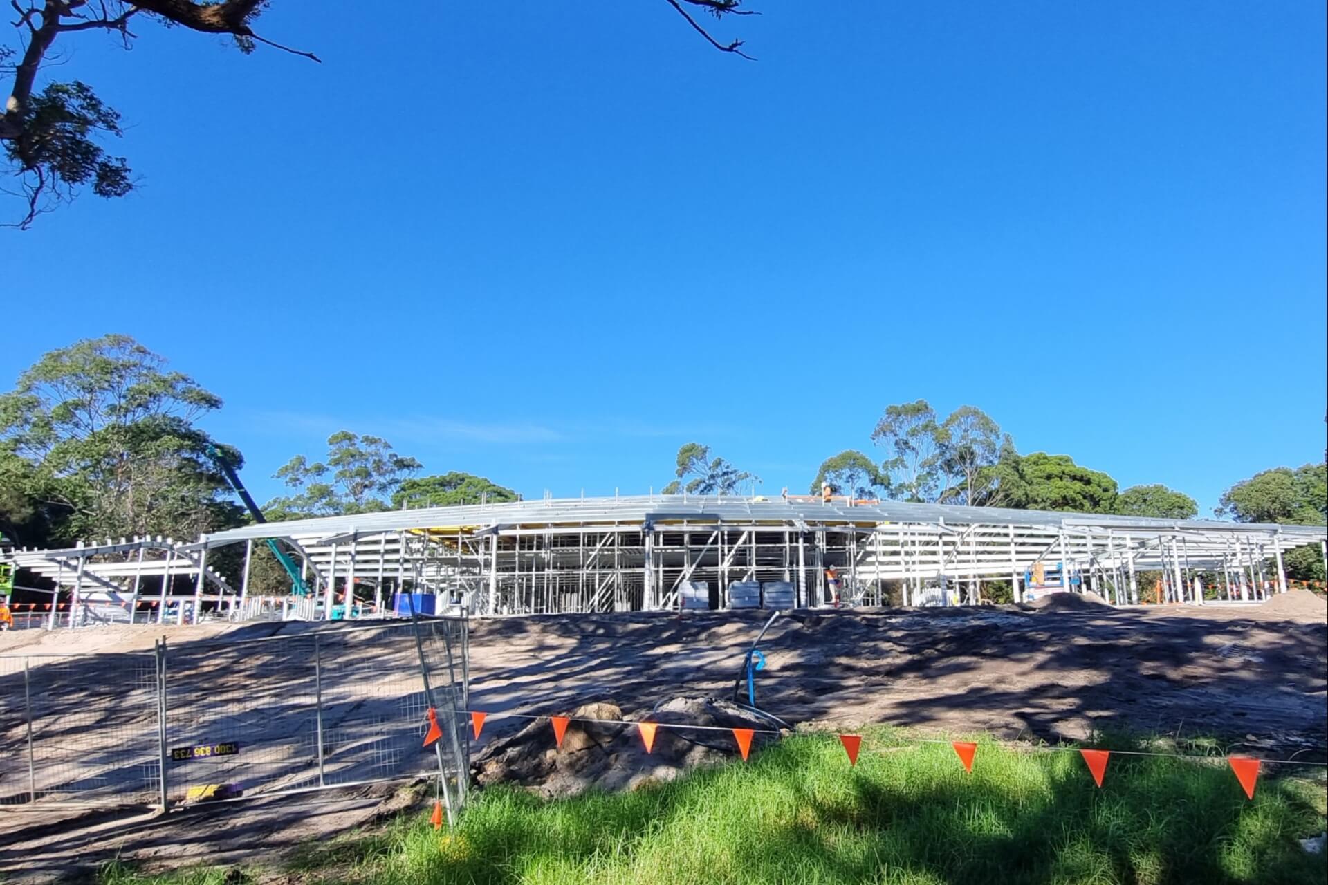 Structural frame for a large single-storey building at a construction site, surrounded by trees and under a bright blue sky
