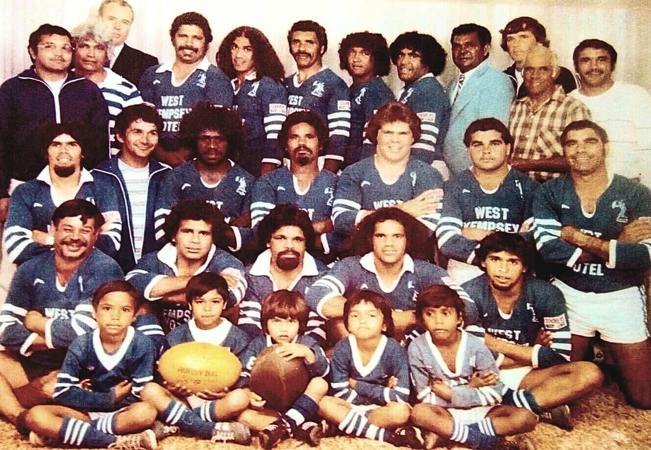 Group photo of the Ngaku Warriors rugby team, 1982. The group is arranged in four rows: men standing in the back row, kneeling in the second row, seated in the third row, and five young children seated in the front. Two of the children are holding rugby balls. Most team members are wearing blue long-sleeved jerseys, white pants, and blue knee-high socks.