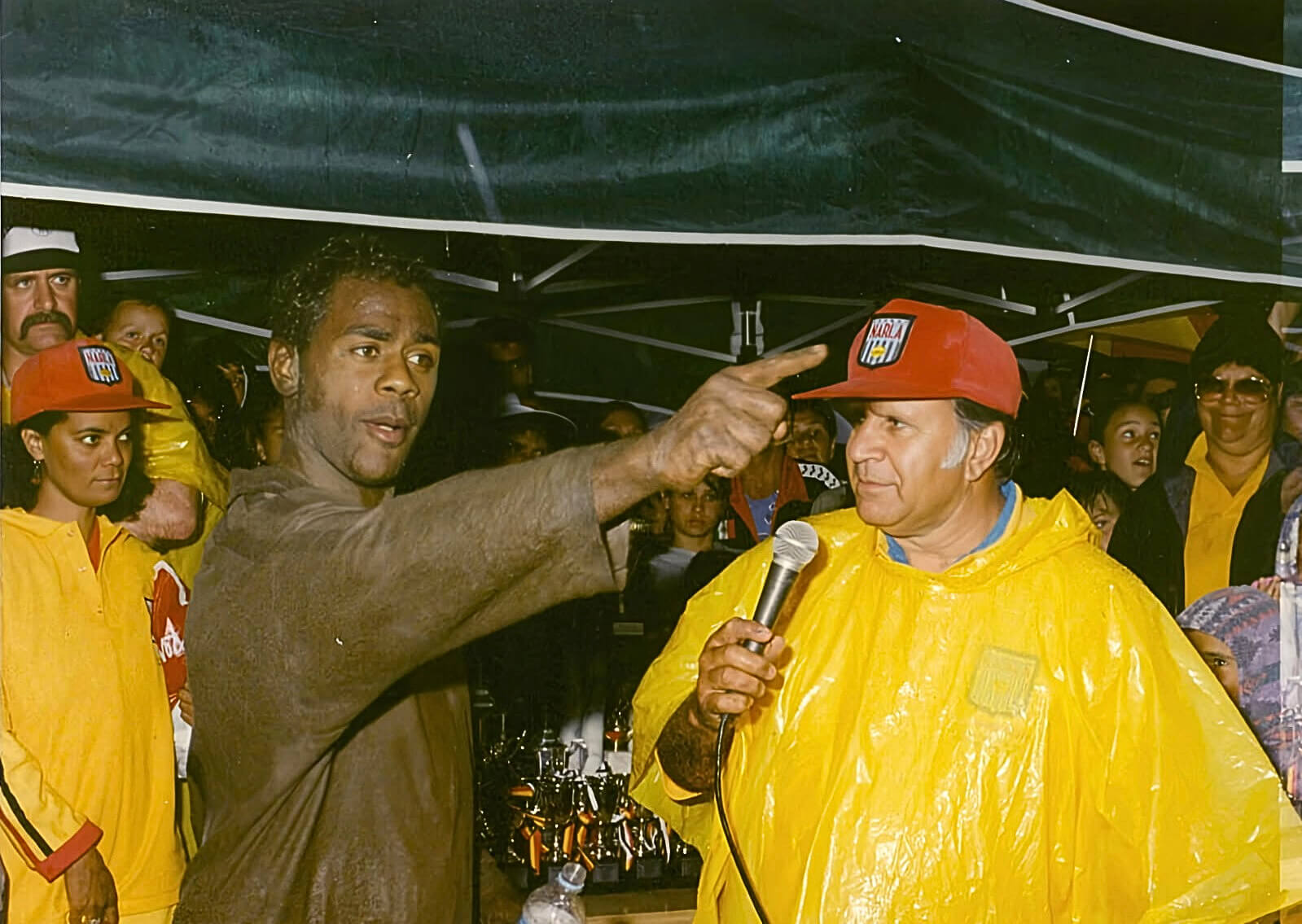 Photo of Shane Phillips enjoying the Knockout, date unknown. He is pointing outward with his finger, while standing next to a male interviewer wearing a cap and a yellow raincoat, who holds a microphone. Behind them, a crowd of people fills the background, capturing the lively atmosphere of the event.
