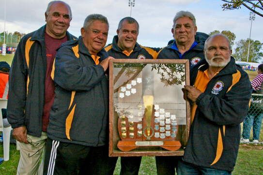 Group photo of Koori Knockout co-founders (L–R) Bob Morgan, Bill Kennedy, Dan Rose, Vic Wright, and Bob Smith in Woy Woy, NSW, 2010. They are smiling at the camera, wearing green and gold team jackets, and proudly holding the original Koori Knockout trophy.