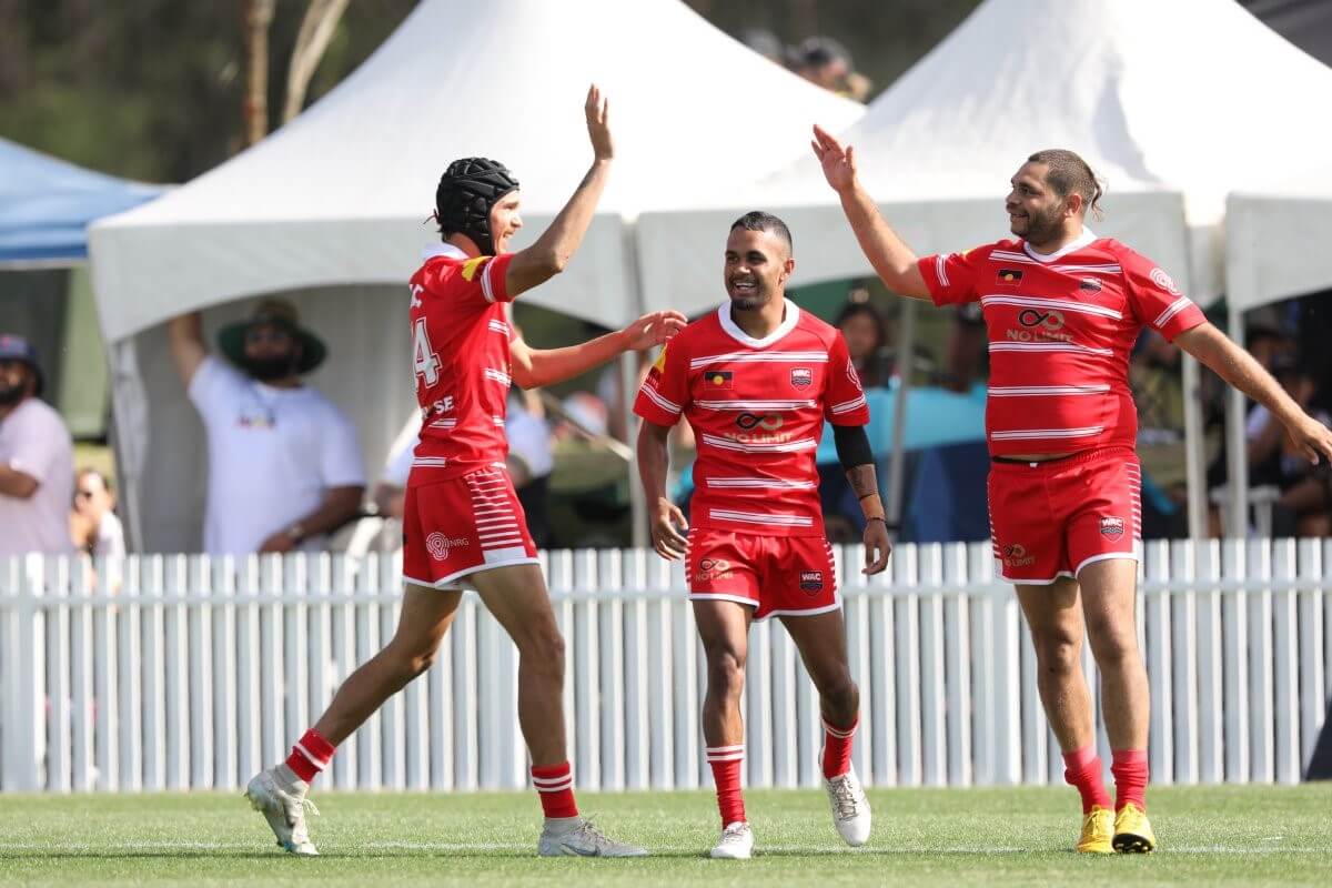 Photo of Christopher Woodbridge, Troyden Dixon, and Dillon Walford from Walgett Aboriginal Connection at the Koori Knockout in Tuggerah, 2023. They are on a rugby field, all wearing matching red rugby T-shirts, shorts, and red socks. Two of them are about to high-five, smiling joyfully, capturing a moment of celebration and team spirit.