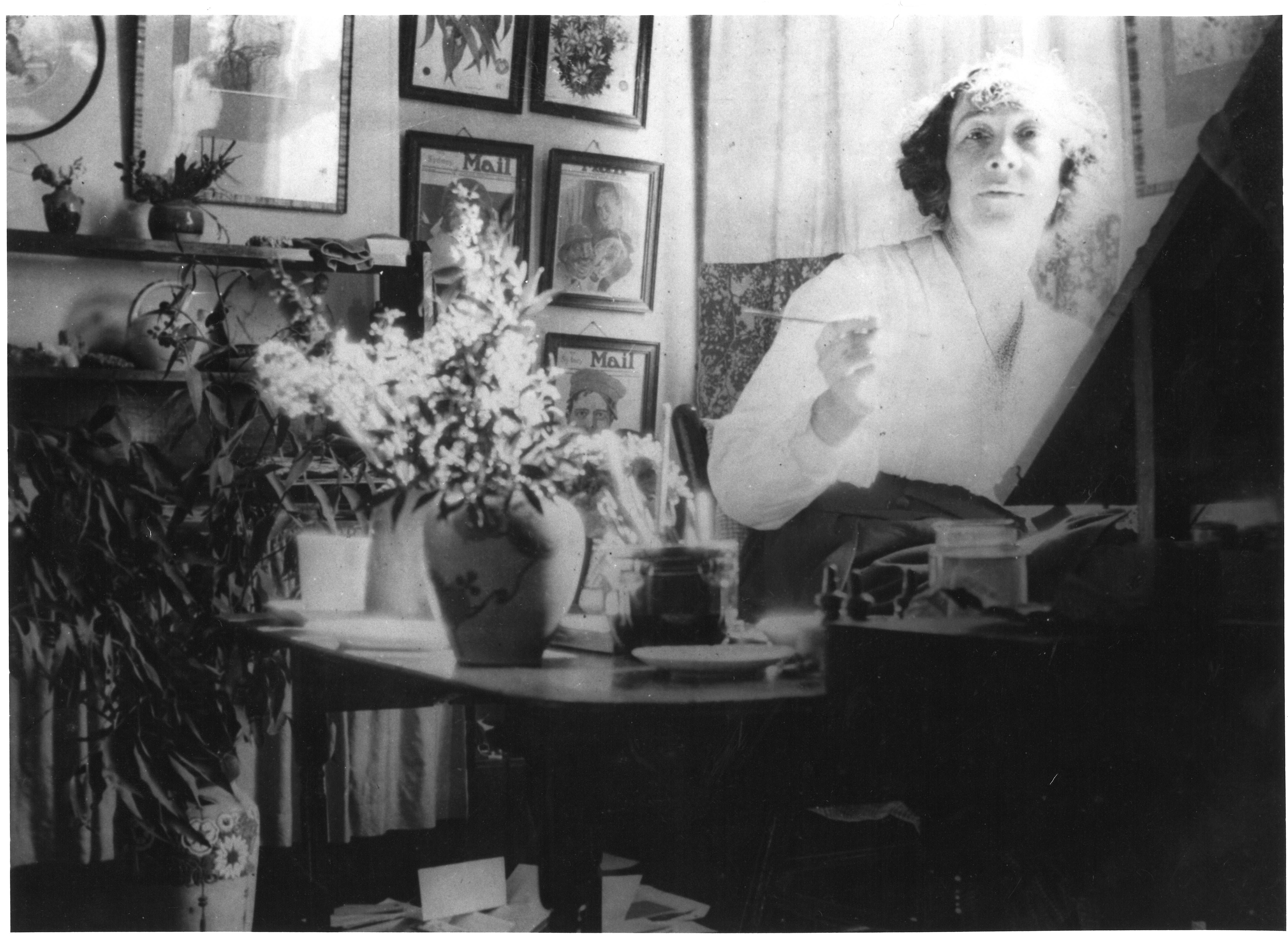 Black and white photograph of May Gibbs working at her rented studio on Bridge St in Sydney. May is seated behind an easel, looking to camera holding a paintbrush. On a table next to her is a vase of native foliage and a series of her artworks for the Daily Mail are framed on the wall behind her.