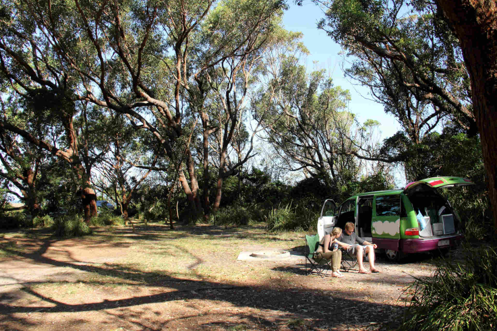 Two people sit in camp chairs beside a van and a fire pit, in a tree-lined clearing with glimpses of the ocean visible through the vegetation