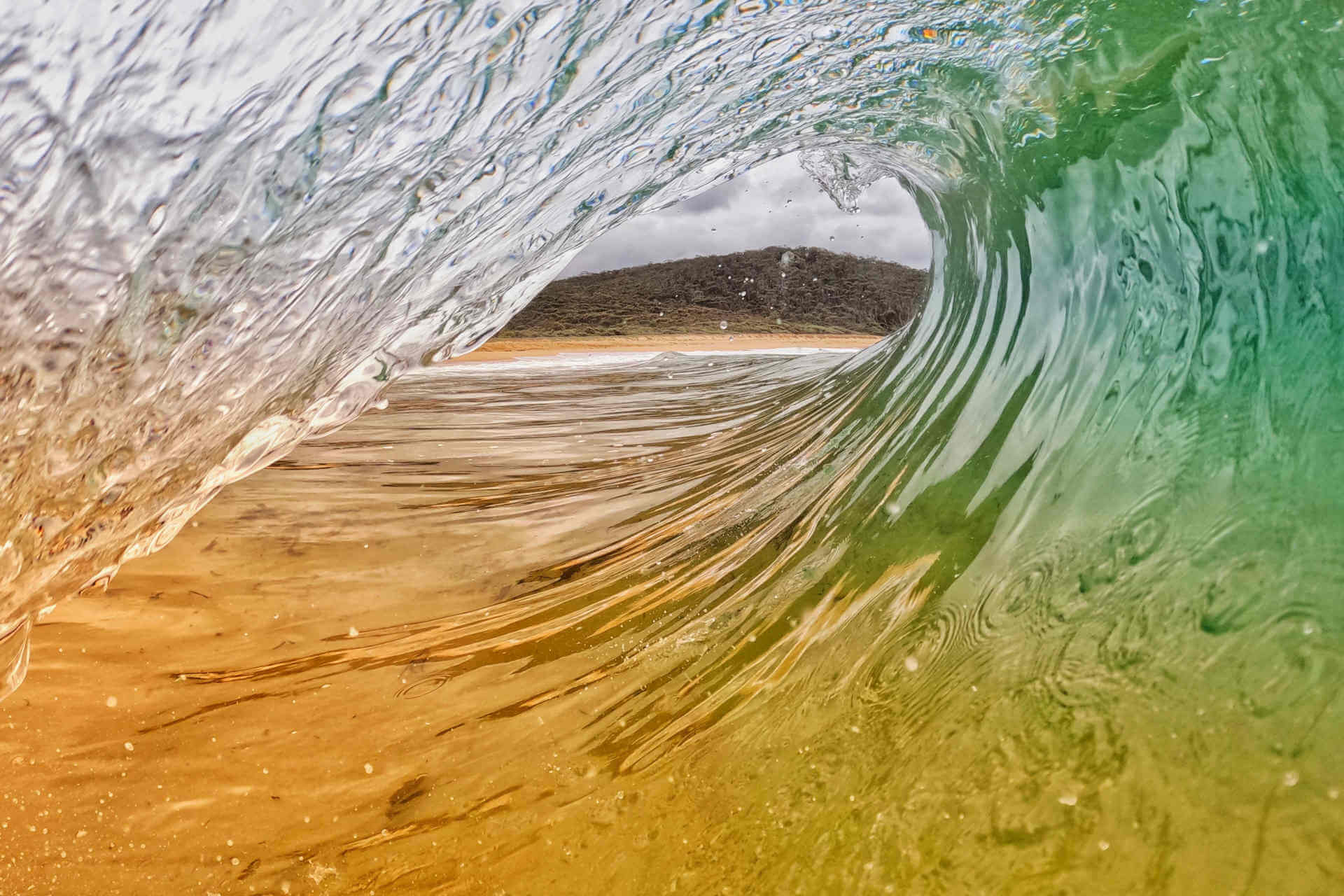 A photograph taken from within the curve of a clear and aqua-green wave, with a thickly treed hill visible on the shore, framed all around by the curvature of the water