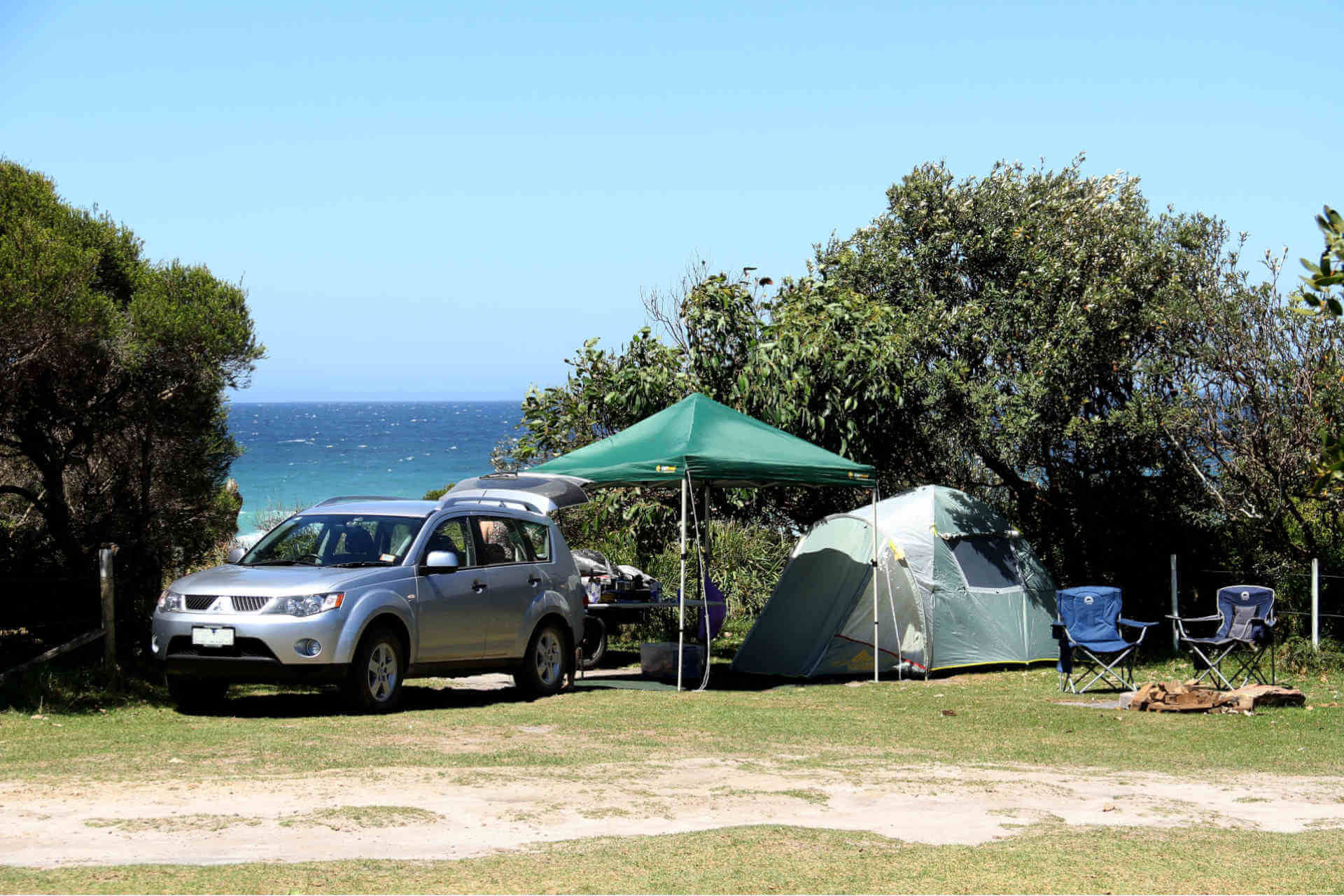 A tent, 2 camp chairs and a gazebo set up behind a car in a green clearing, with the ocean visible through a break in the treeline