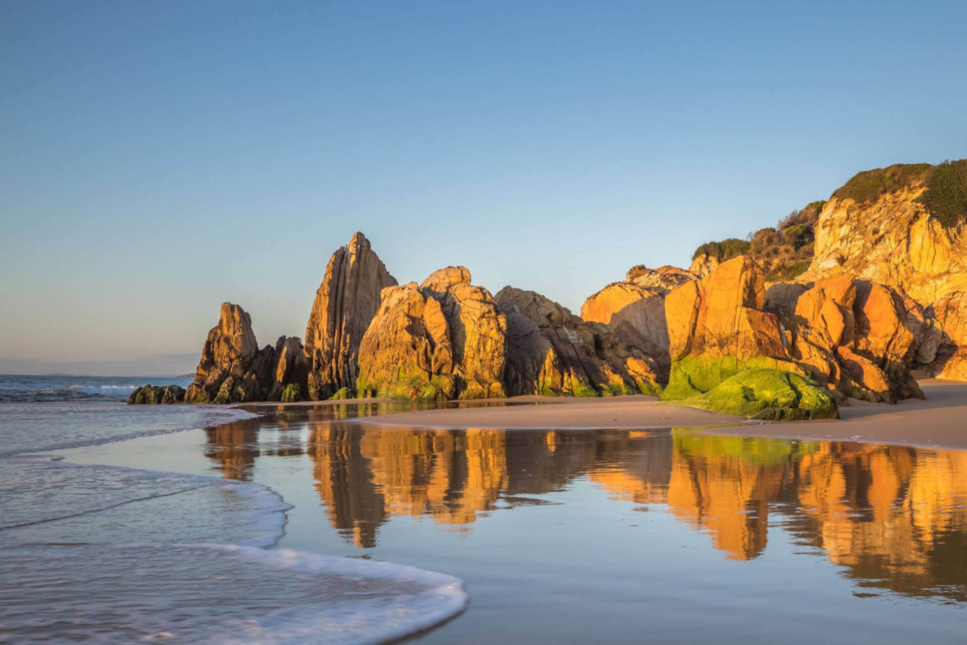 Large headland boulders are glowing golden in a gentle light, with the picture reflected in the calm water along the seashore