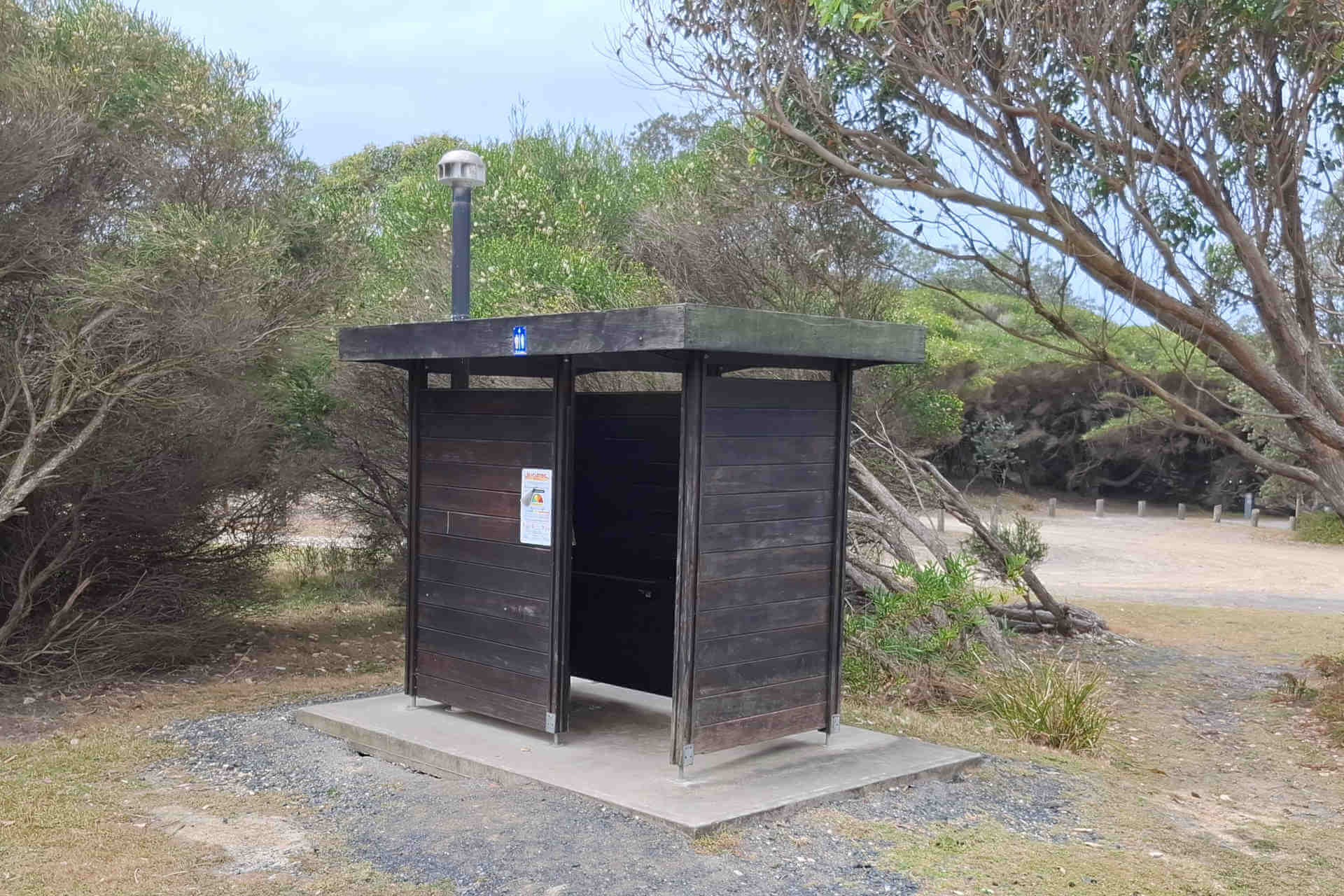 A small, single wooden hut toilet, with coastal vegetation and a cleared road area behind it