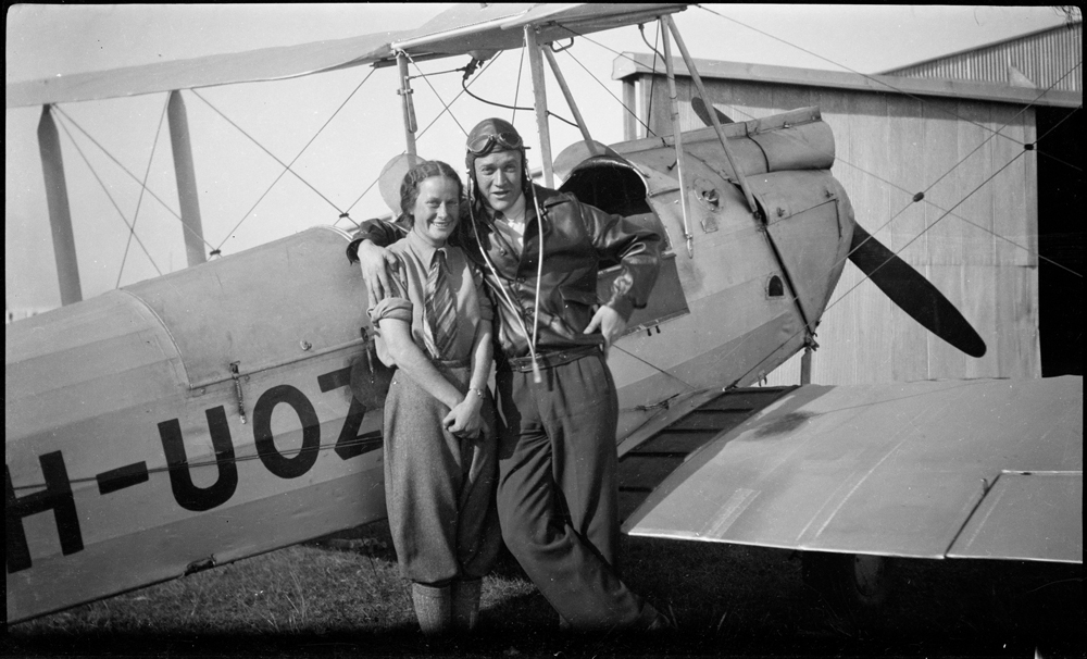 Black and white image of a man and a woman standing in front of an old aeroplane with ‘H-UOZ’ on the side.