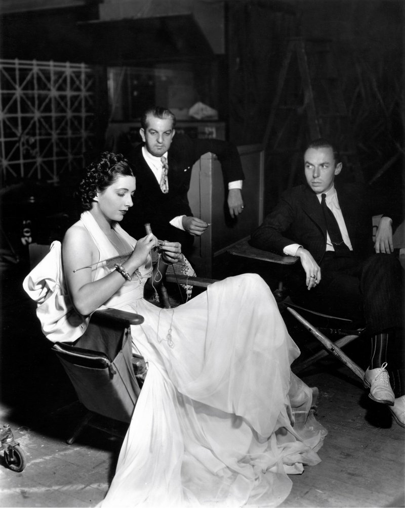 Black and white candid photo from the set of Another Dawn (1937), featuring actress Kay Francis and costume designer Orry-Kelly seated together, with another man beside them. Kay Francis is focused on knitting, wearing a formal long white dress. Orry-Kelly, seated in the middle, and the other man are both dressed in suits and ties, watching Kay with interest.