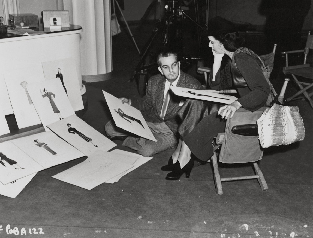 Black and white photo of Orry-Kelly in a design studio, seated on the floor alongside a woman sitting on a chair. Both are holding large fashion design prints and looking at them intently. Numerous other design drawings are scattered across the floor, creating a creative and busy atmosphere in the studio.