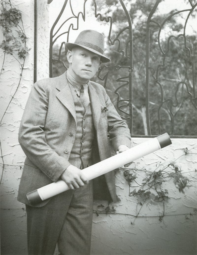 Black and white photo of Paul Sorensen dressed in a suit and hat, stands against a wall covered with climbing plants, holding a large rolled-up document or blueprint. Behind him is an ornate window with metalwork designs and trees visible through the glass.