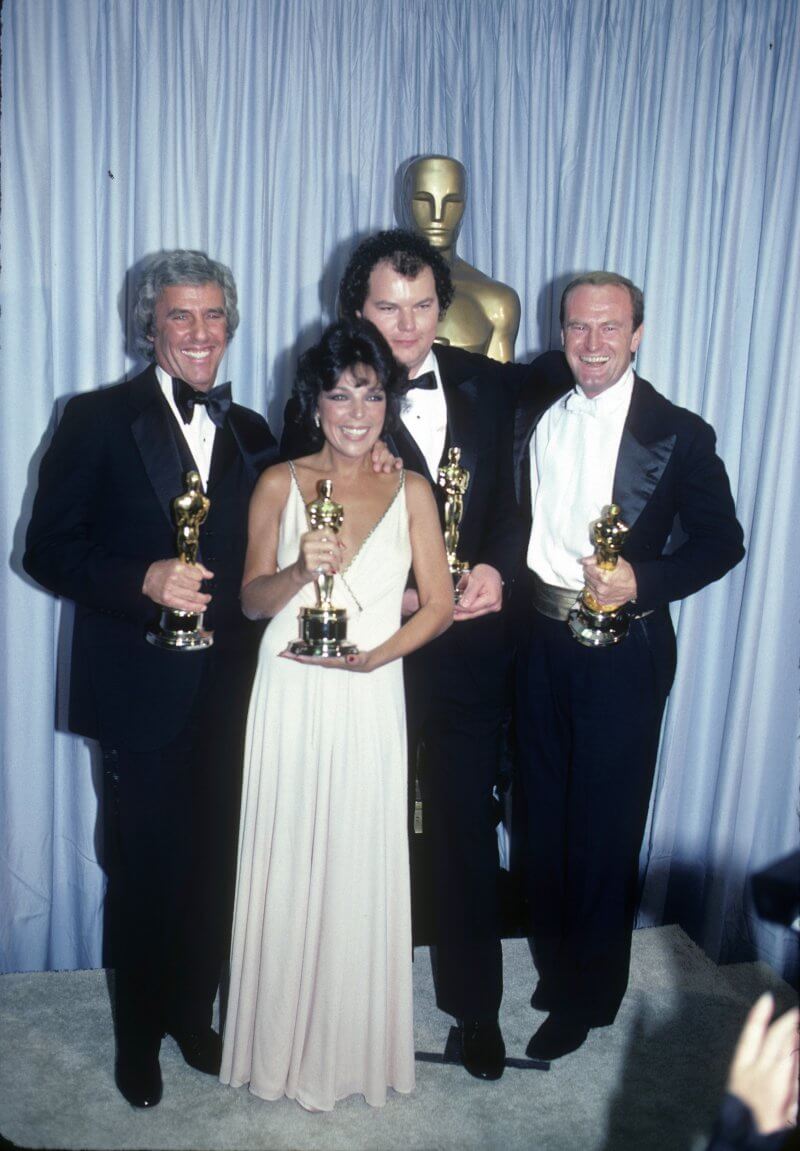 Backstage at the 54th Annual Academy Awards on March 29, 1982. From left to right: Burt Bacharach, Carole Bayer Sager, Christopher Cross, and Peter Allen, winners of Best Original Song for 'Arthur's Theme (Best That You Can Do).' The men wear black suits, and Carole wears a long white sleeveless dress. All are smiling at the camera, holding their Oscar trophies.