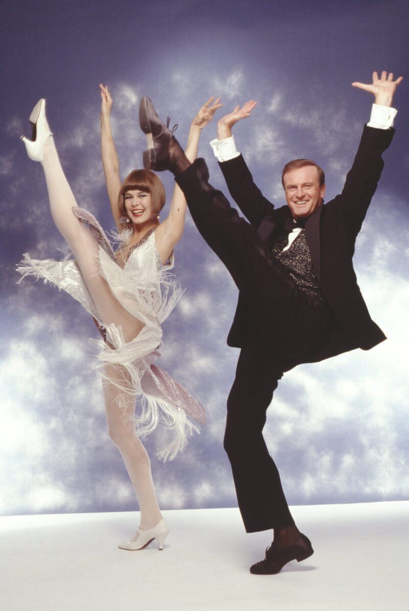 Peter Allen and Colleen Dunn performing in the Broadway musical 'Legs Diamond' in September 1988. Peter wears a black three-piece suit and Colleen a white shimmery sleeveless dress. Both are mid-kick with their left legs raised high, arms up, smiling brightly at the camera.