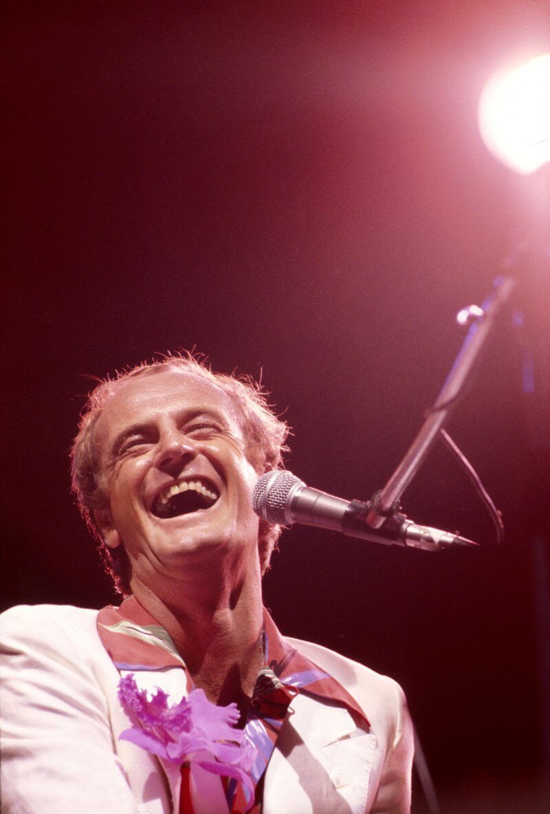 Peter Allen performing on stage in New York, 1977. He is laughing joyfully with his mouth wide open and eyes closed, wearing a white suit with a red shirt. A microphone stands in front of him.