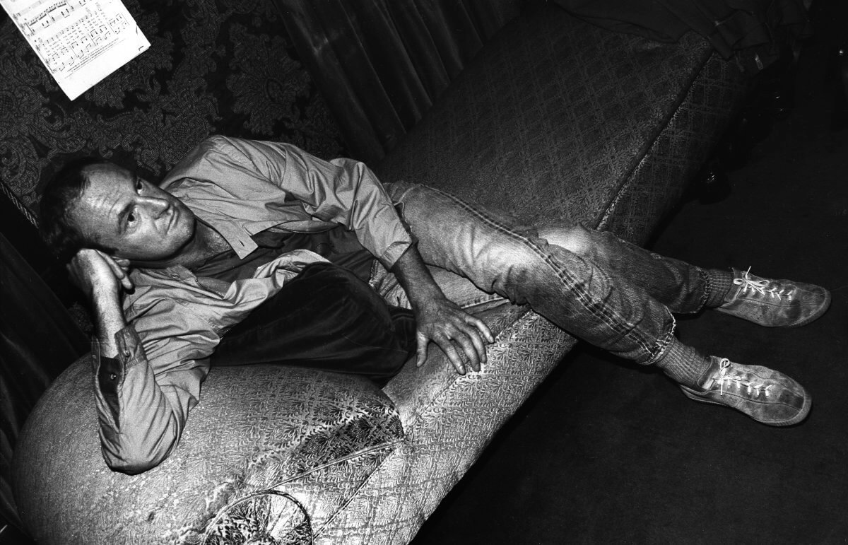 Black and white portrait of Oscar-winning entertainer Peter Allen sitting on a couch, resting his head on one hand, legs crossed, photographed from a high side angle.