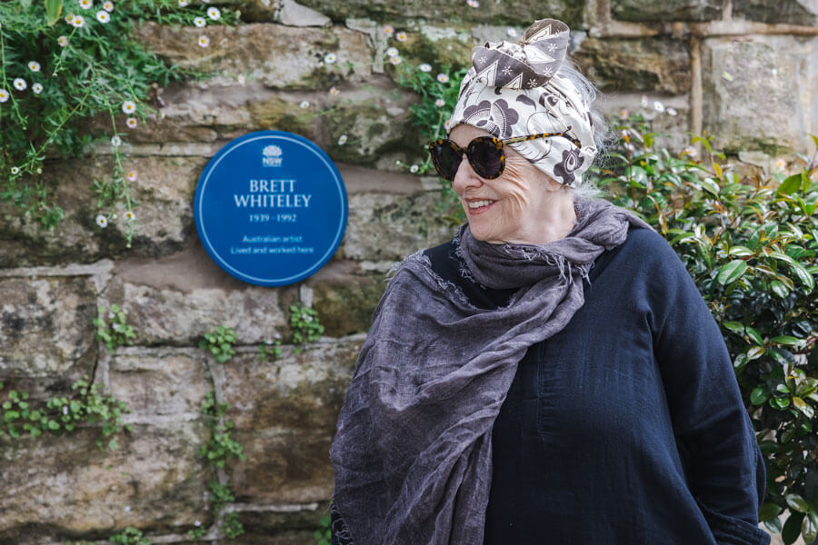 Wendy Whiteley standing in front of her Lavender Bay home, wearing sunglasses and looking to the left. Her hair is wrapped in a grey and white patterned bandana. She wears a dark blue linen long-sleeved top with a grey scarf draped around her neck.
