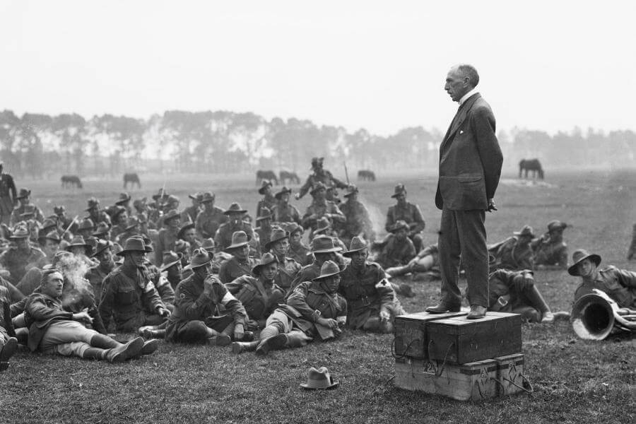 Billy Hughes standing on 4 ammunition boxes, addressing men a part of the 5th Australian Field Ambulance on an open grass field with horses in the background.