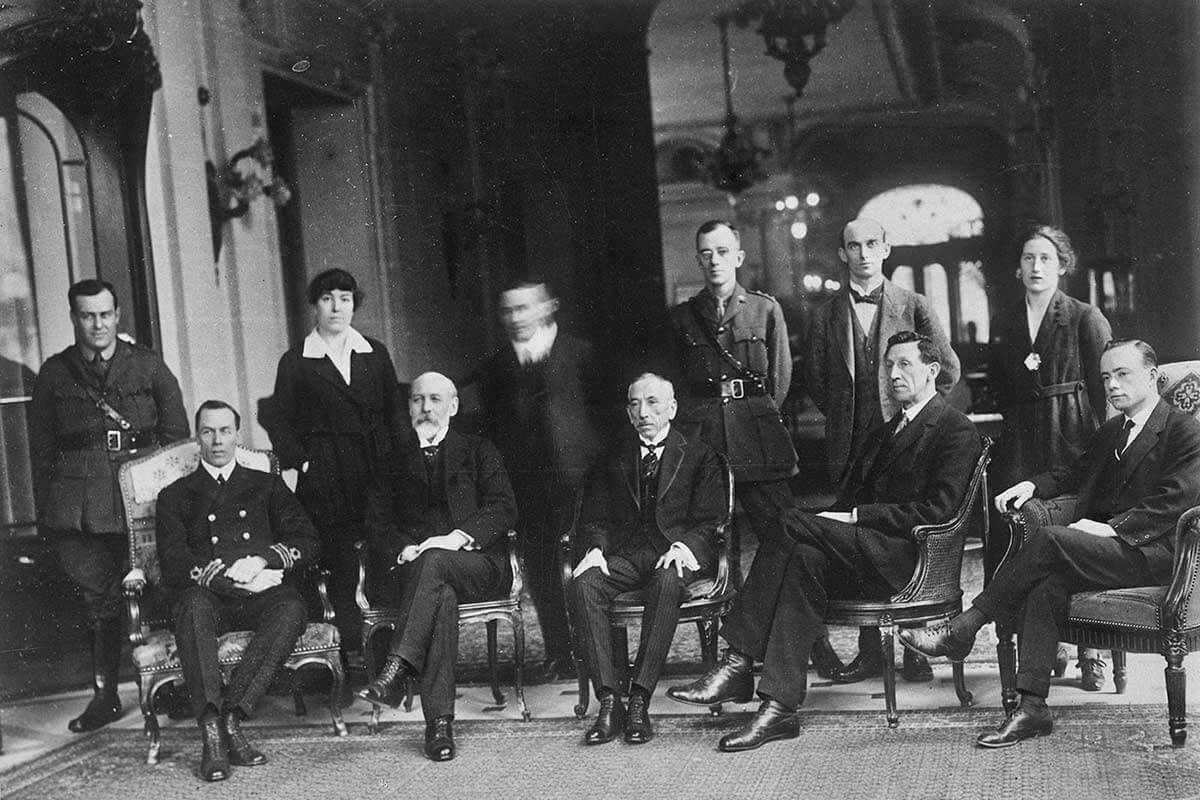 Black and white image of 4 men and 2 women standing behind 5 men sitting on chairs.