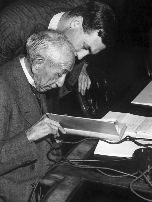 Black and white image of Billy Hughes sitting down accompanied by another man looking over his shoulder, both viewing a document.
