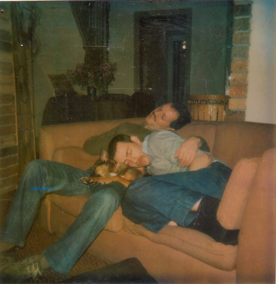 Photo of two men lying close together on a couch, with a dog peacefully sleeping on one of their laps. Both have their eyes closed, and one man has his arms wrapped gently around the other, conveying a sense of comfort and affection.