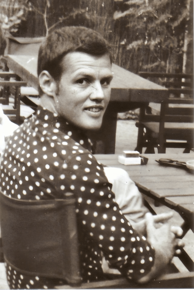 Sepia-toned photo of Bobby Goldsmith sitting on a chair outdoors, with his back to the camera. He turns his head over his shoulder and smiles warmly at the camera. He is wearing a dark shirt with polka dots. A table is beside him, and the setting suggests a relaxed, casual moment in a natural environment.
