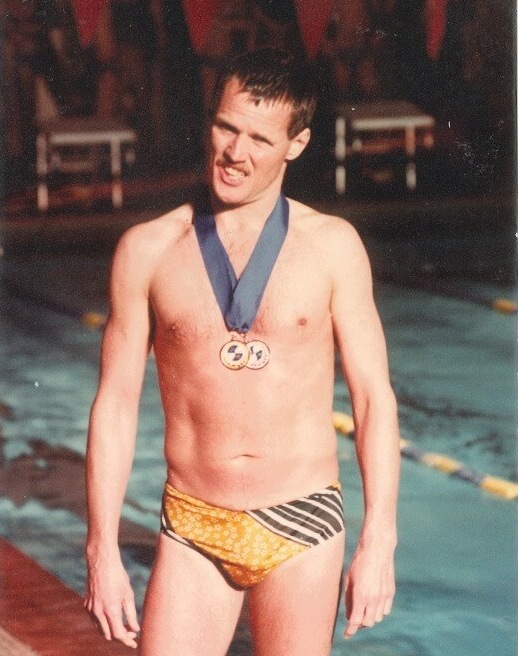 Photo of Bobby Goldsmith standing beside a swimming pool at the Gay Games. He is wearing a yellow and brown patterned swim brief and has two medals hanging around his neck. He is looking directly at the camera with his teeth showing.