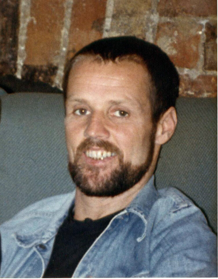 Photo of Bobby Goldsmith sitting in a relaxed pose, wearing a blue denim shirt over a black T-shirt. He is looking at the camera, smiling with his teeth visible. He has a moustache and beard, contributing to his warm and approachable appearance.