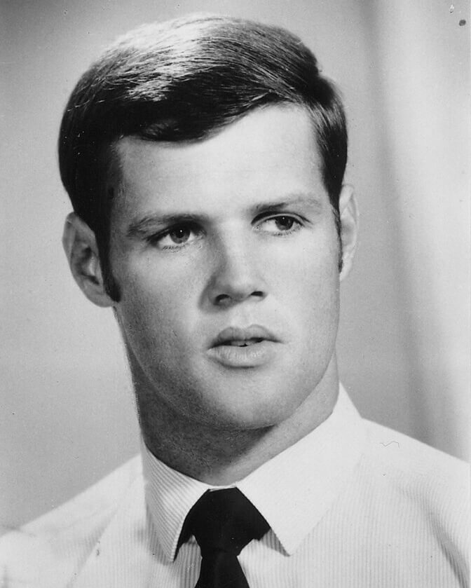 Black and white portrait of Bobby Goldsmith. He is wearing a white shirt with light vertical stripes and a dark tie. He is looking slightly away from the camera into the distance, with a calm and reflective expression.