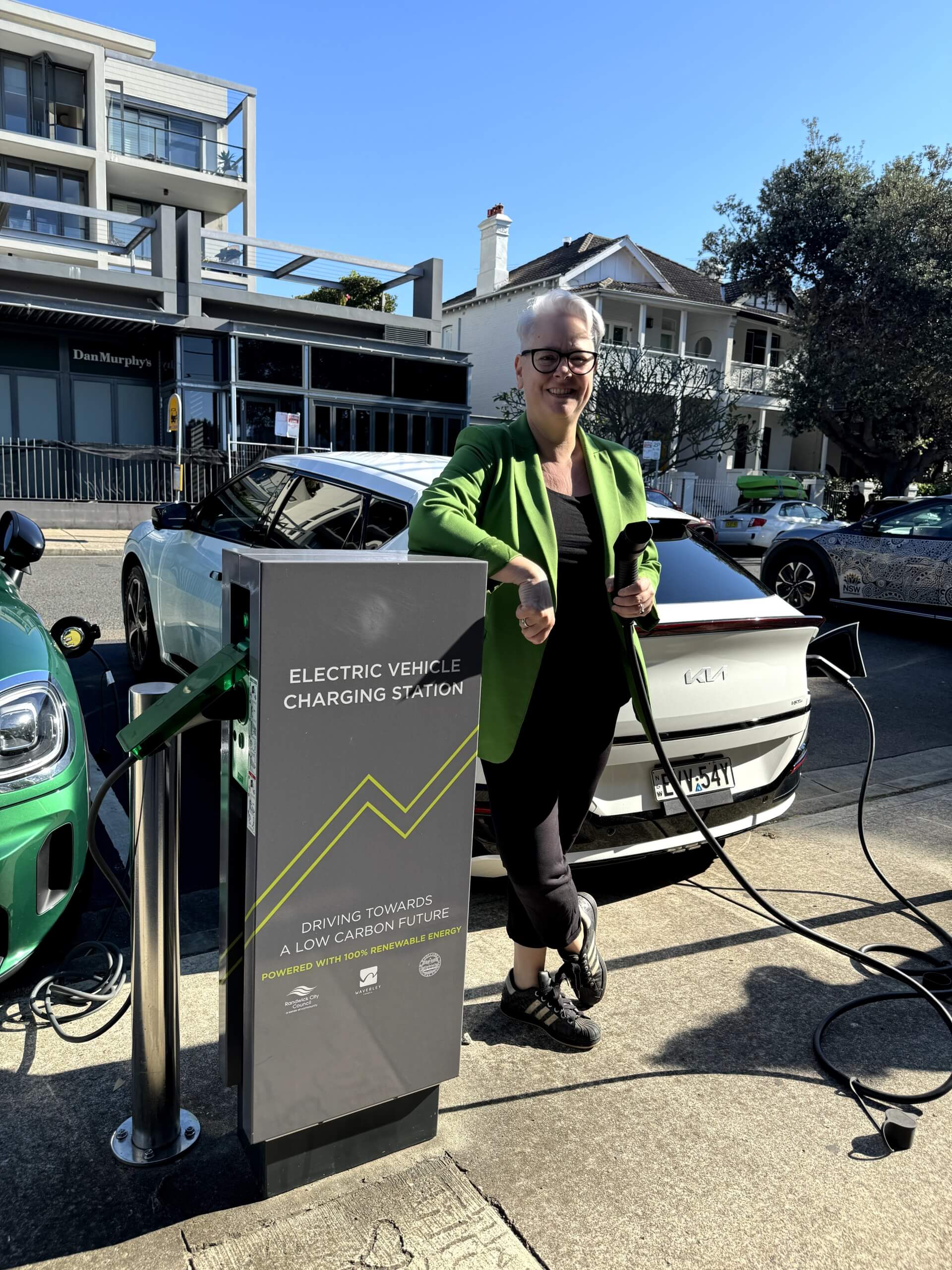 A woman stands next to a tall, grey EV charging station. There are cars and buildings in the background.