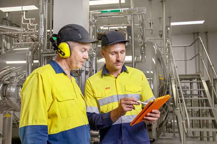 Two men in hi-vis and protective gear stand looking at clipboard, machinery behind them.