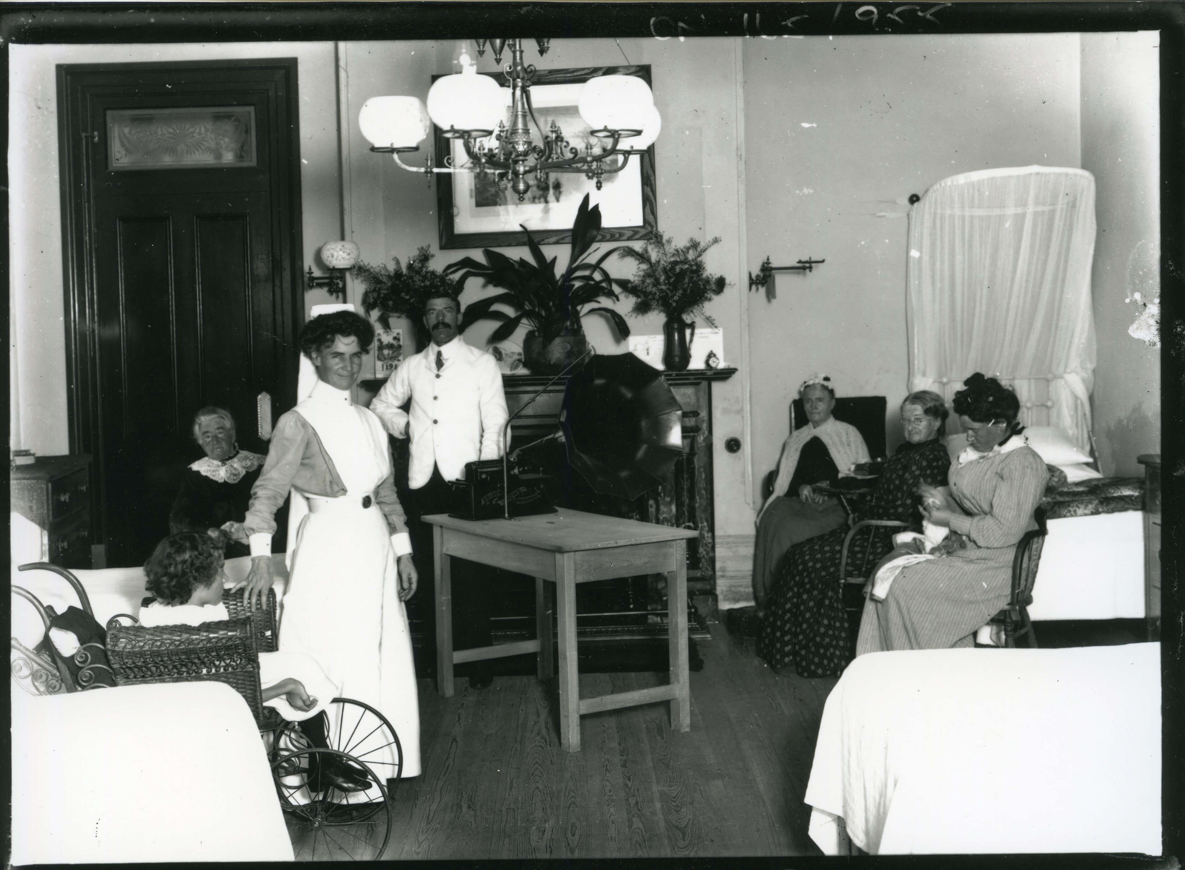 Black and white photo of a room. Standing in the room is a woman wearing a white nurse’s outfit and a man wearing a white suit. Patients are sitting on a bed nearby. In the middle of the room is a gramophone.