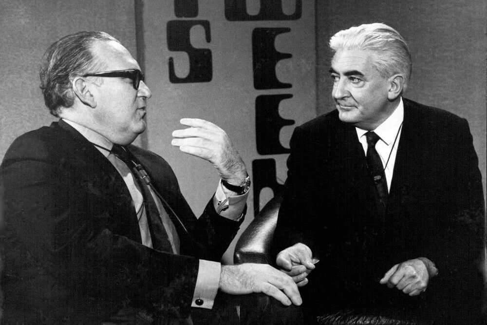 A black and white photograph of 2 men talking to each other, sitting in armchairs on a TV set