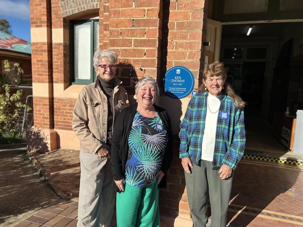 Three women pose in front of brick building with a blue plaque