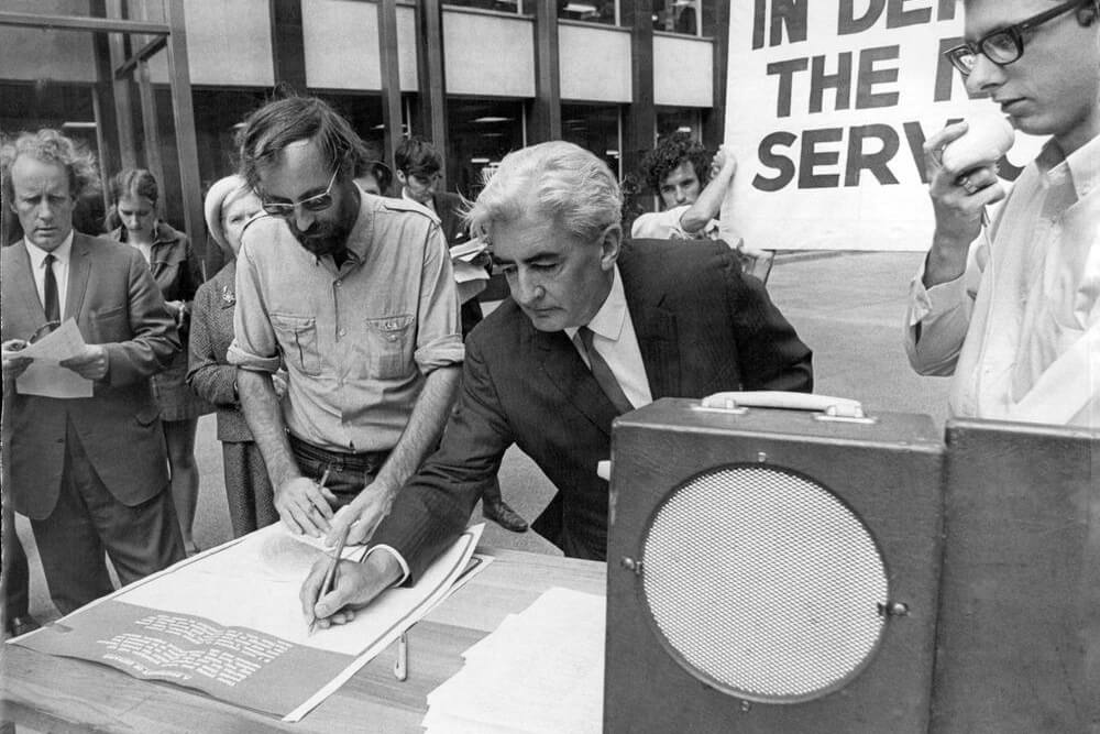Black and white photograph of a man signing a document, with a crowd of protesters in the background